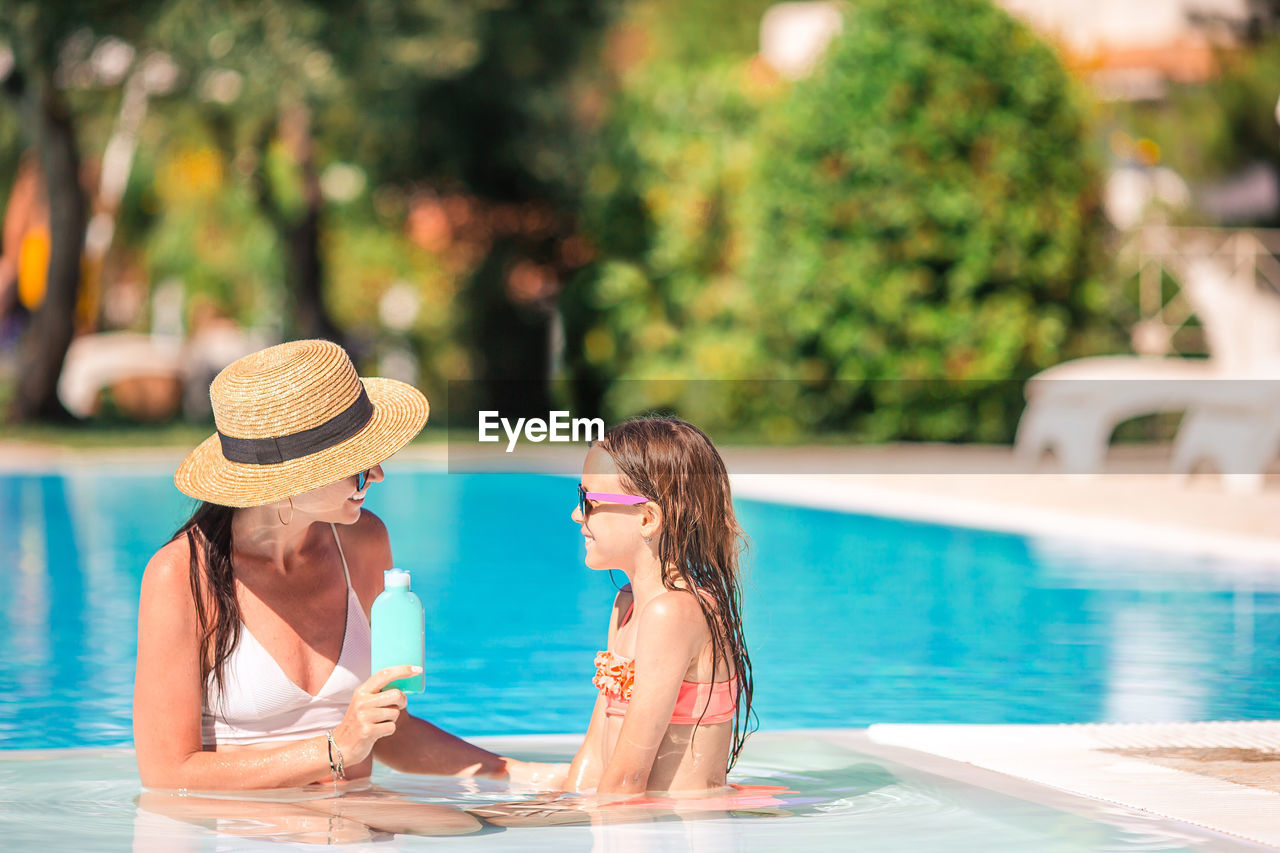 Smiling mother and daughter wearing hat sitting by swimming pool