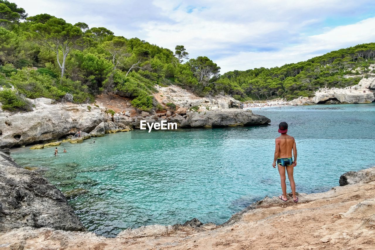 Rear view of shirtless boy looking at bay of water against sky