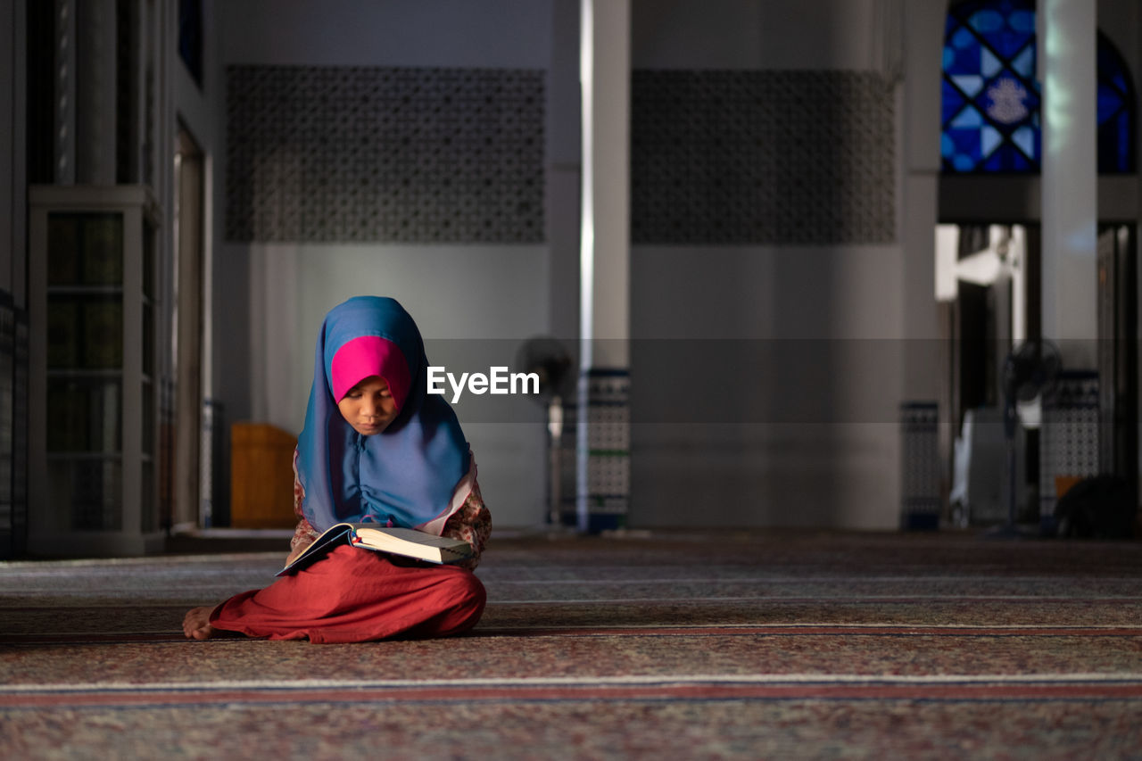 Girl in hijab reading book while sitting at mosque