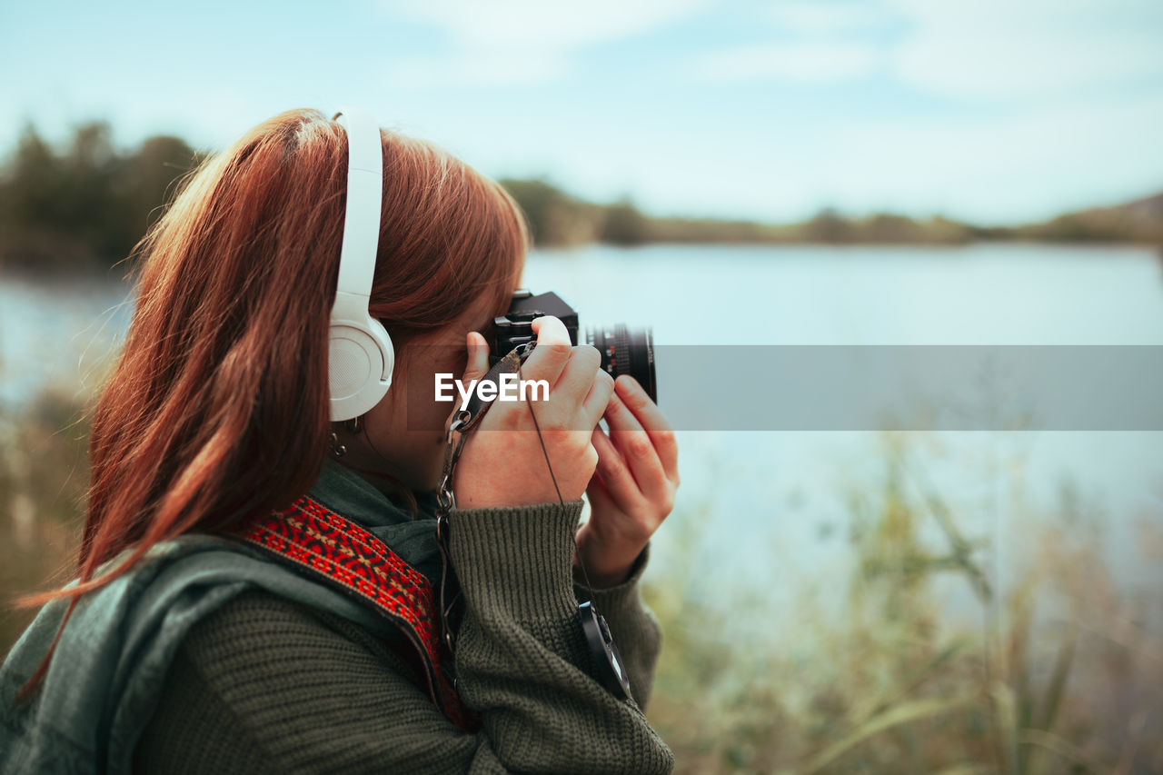 Side view of woman photographing by lake against sky