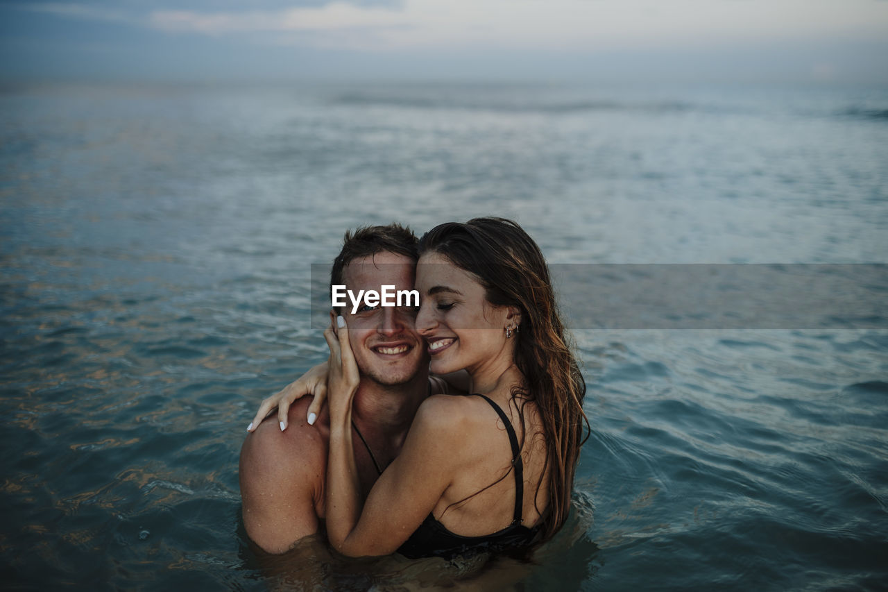 Smiling couple embracing while standing in water at beach during sunset