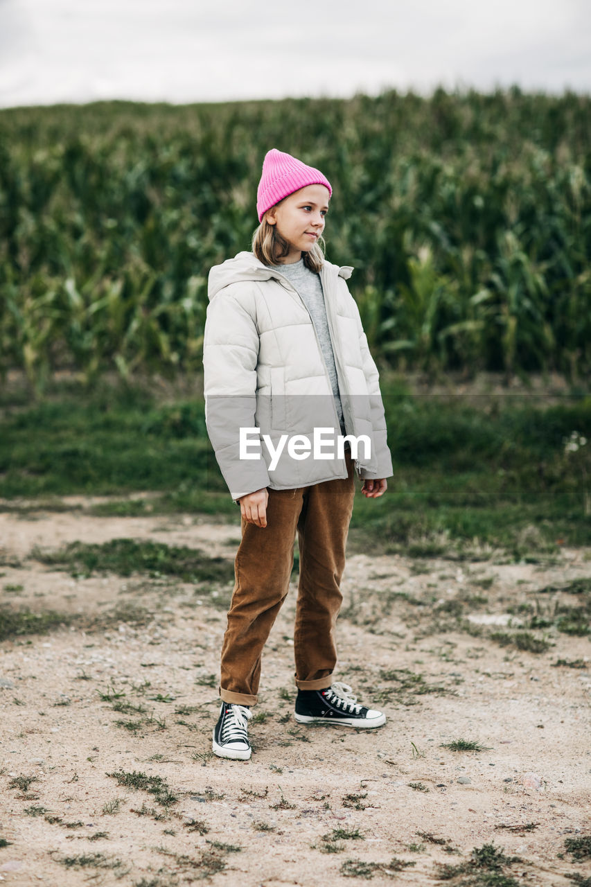 A teenage girl in a stylish image and a pink cap stands against the background of a corn field