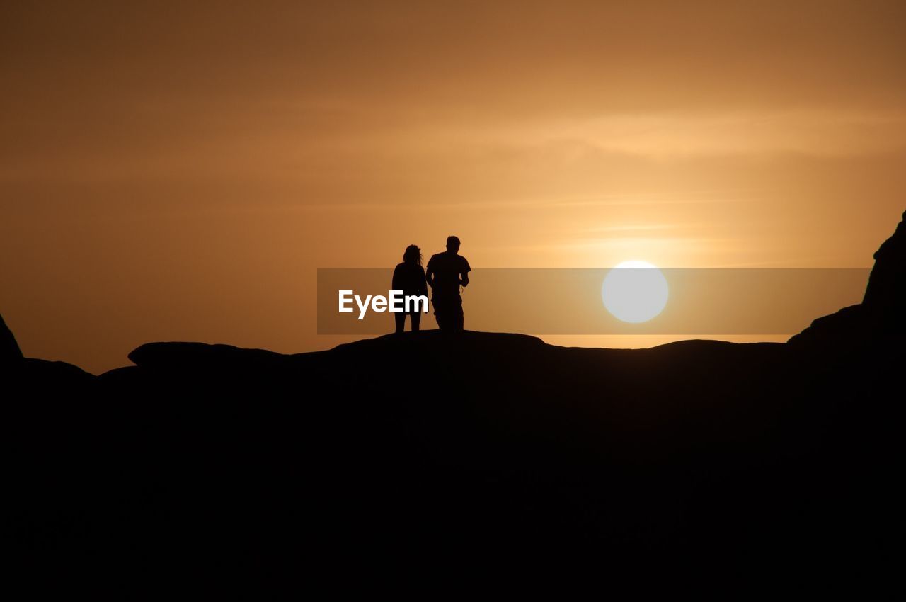 Silhouette people standing on rock formation against sunset sky