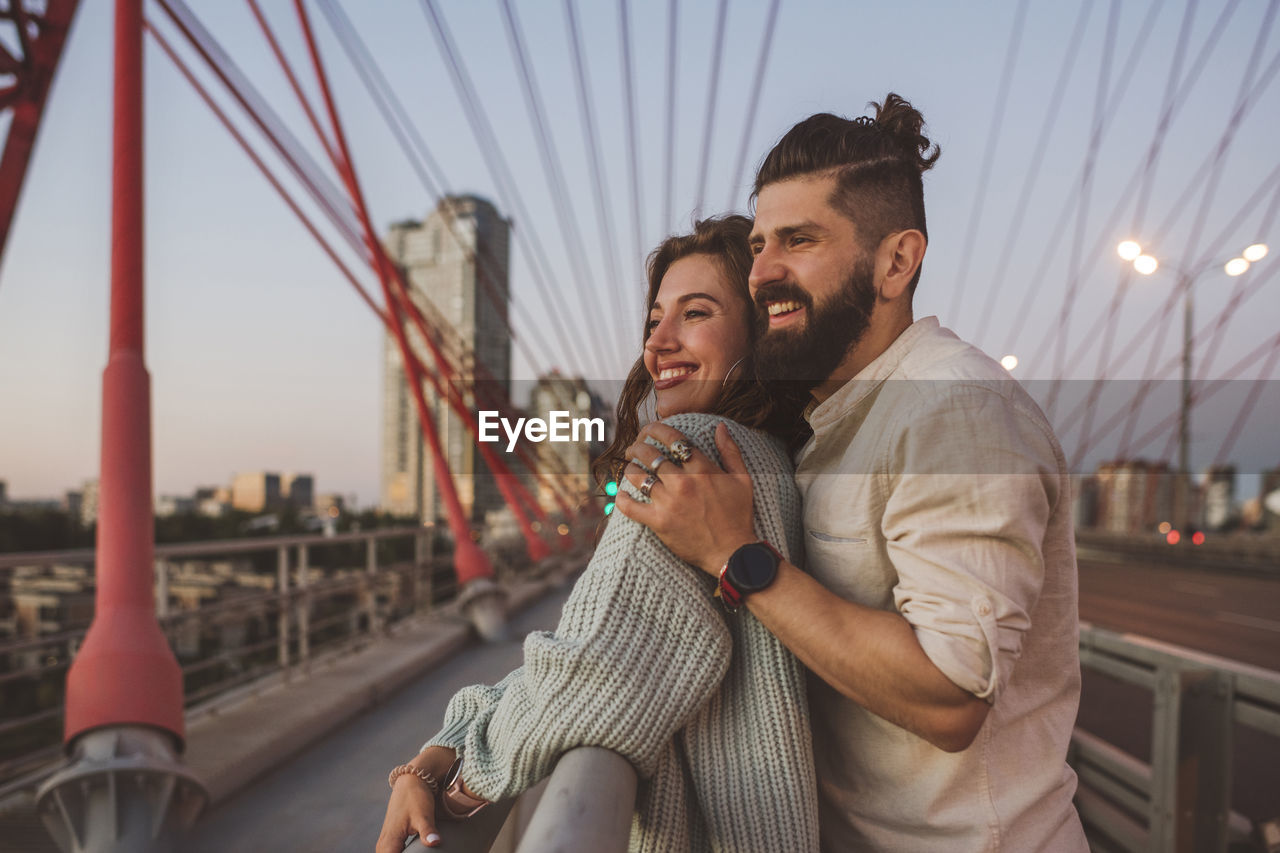 Cheerful boyfriend and girlfriend standing on bridge in city during sunset
