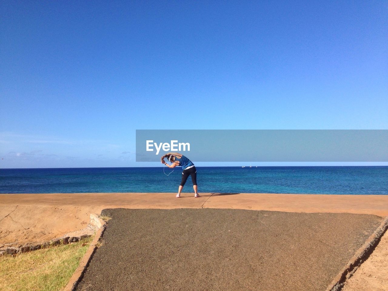 Rear view of woman exercising by beach against clear blue sky