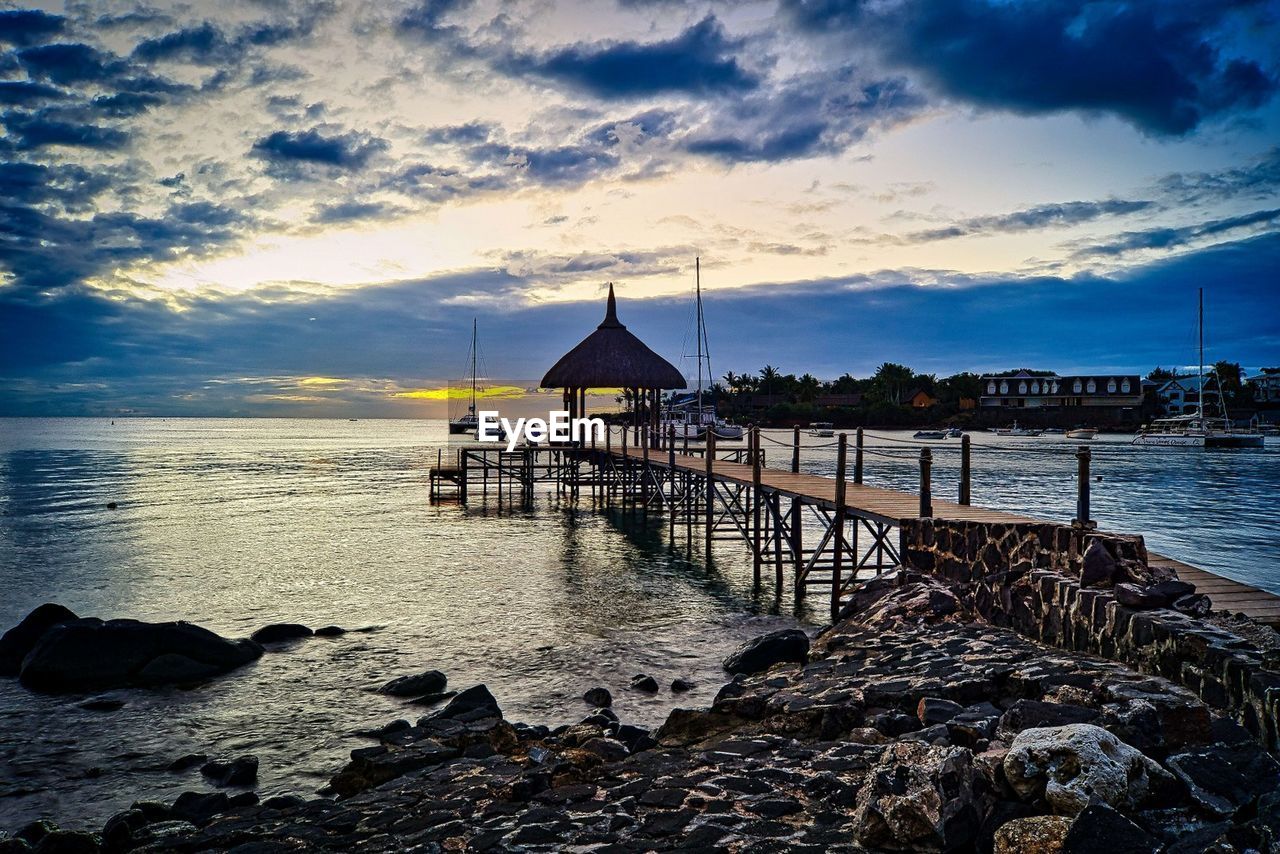 water, sky, sea, ocean, shore, beach, cloud, coast, architecture, land, horizon, nature, sunset, built structure, dusk, wave, beauty in nature, scenics - nature, pier, evening, body of water, travel destinations, tranquility, reflection, tranquil scene, building exterior, bay, holiday, hut, travel, vacation, trip, building, rock, outdoors, idyllic, horizon over water, stilt house, no people, coastline, seascape, sand, jetty, environment, wood, tourism, house, landscape, blue, fishing, transportation, sunlight