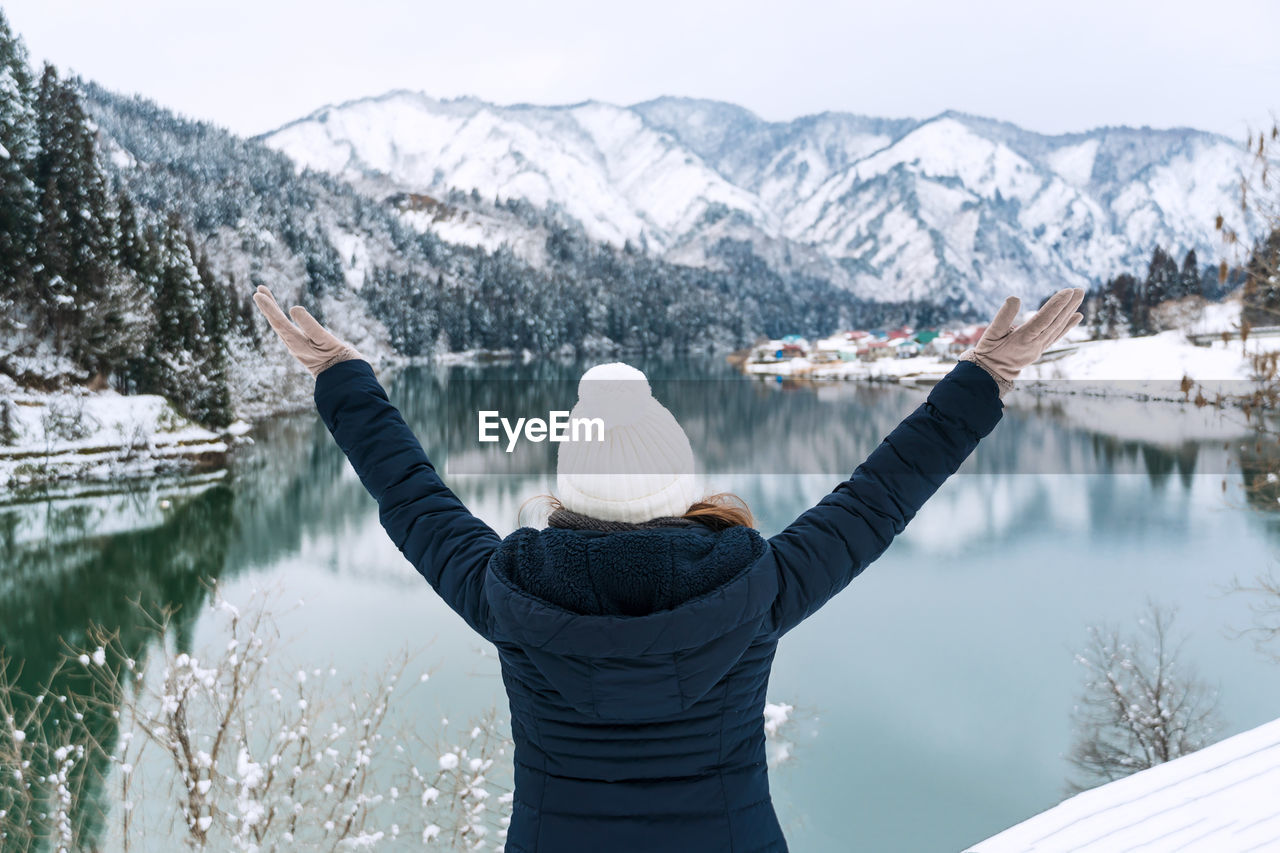 Rear view of woman with arms raised looking at lake against snowcapped mountain