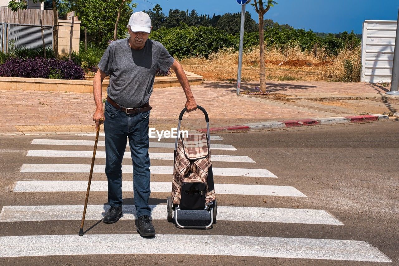full length, road, one person, adult, men, transportation, pedestrian crossing, asphalt, hat, day, city, casual clothing, road marking, architecture, nature, sunlight, occupation, walking, street, crossing, working, person, outdoors, zebra crossing, holding, sign, clothing, motion