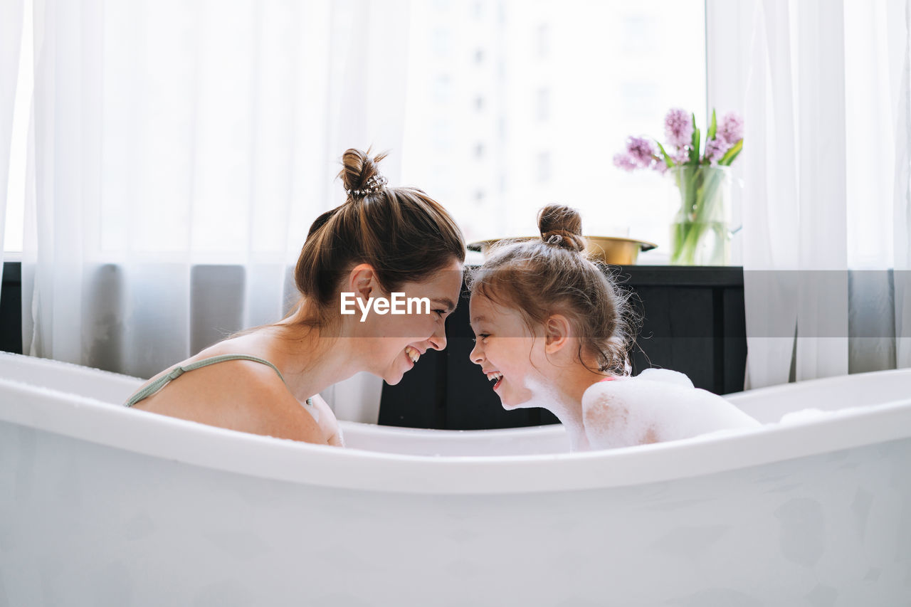 Young woman with little tween girl daughter in pajamas having fun in the bath with foam at home