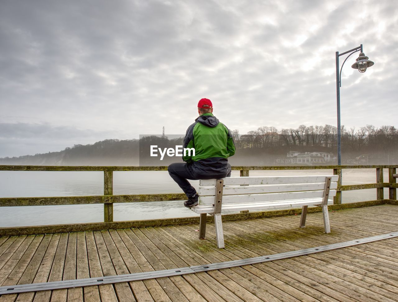 Rear view of man standing by railing against smooth baltic sea during rainy season