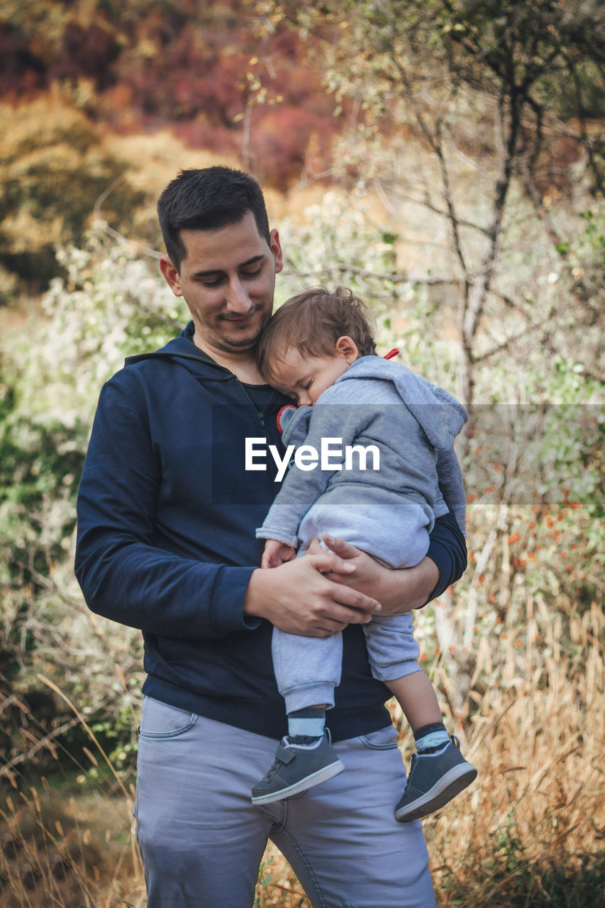 Father carrying sleeping son while standing in forest