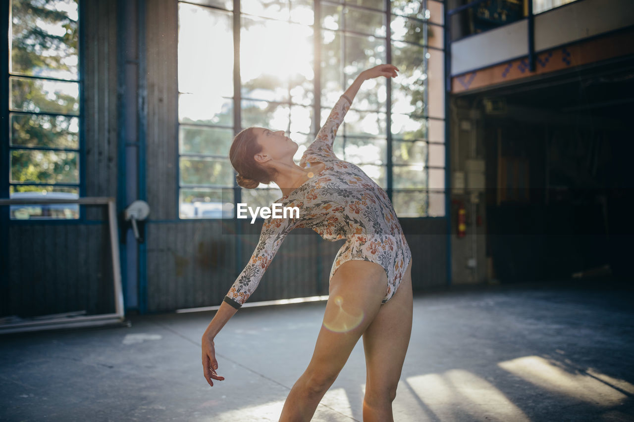 Side view of young graceful ballerina looking up in colorful bodysuit dancing training in light studio