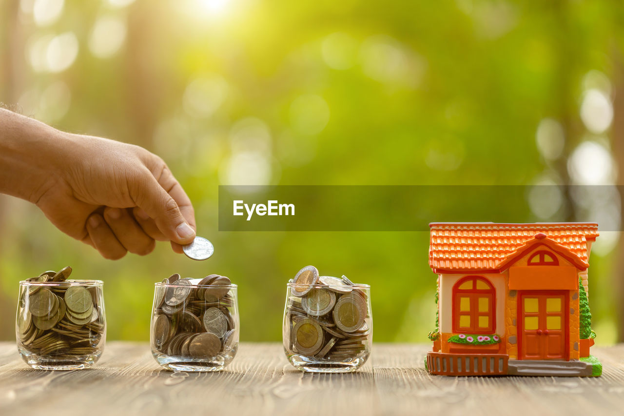 Cropped image of hand putting coins in glass by model home on table