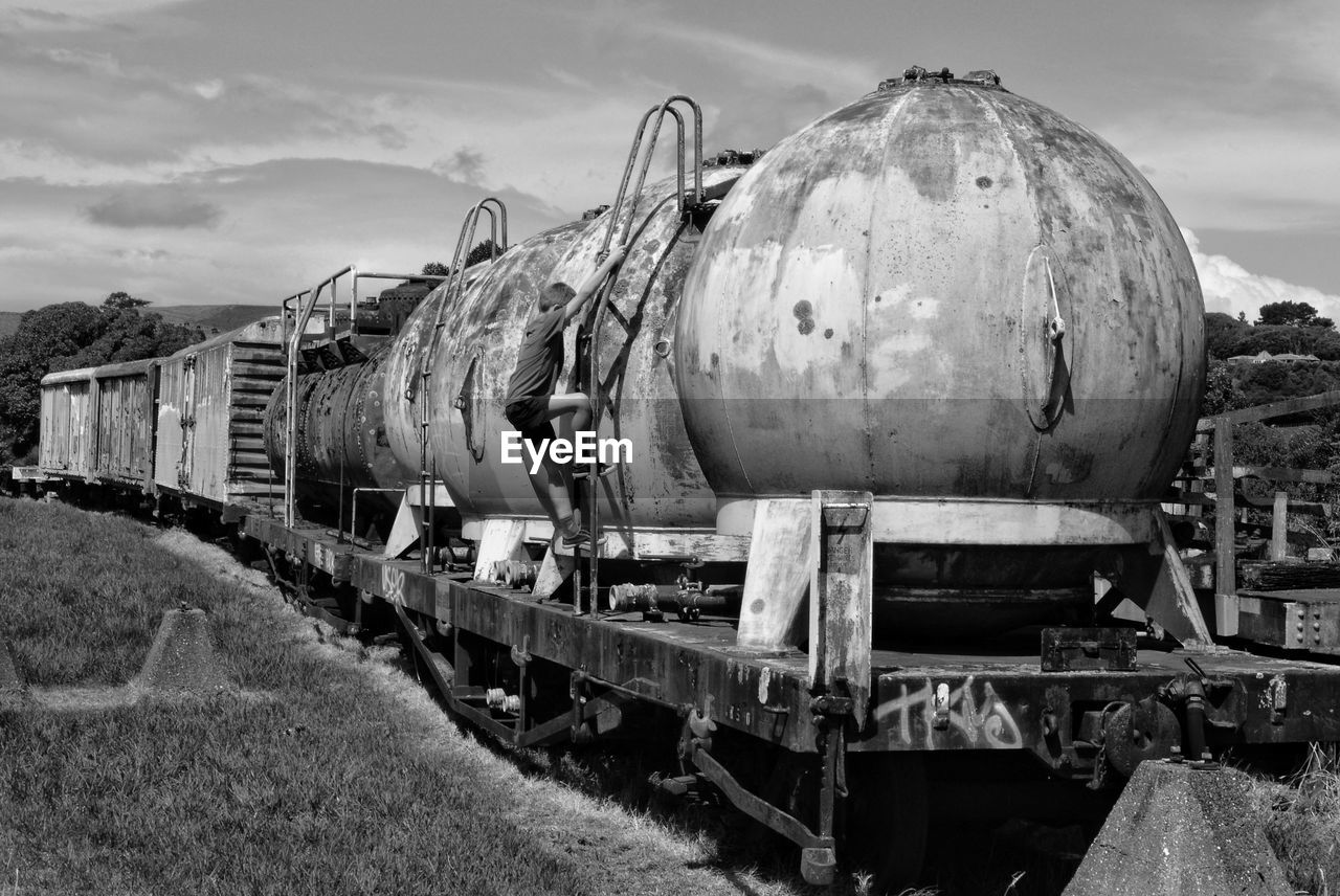 Boy climbing on abandoned freight train on railroad track against sky