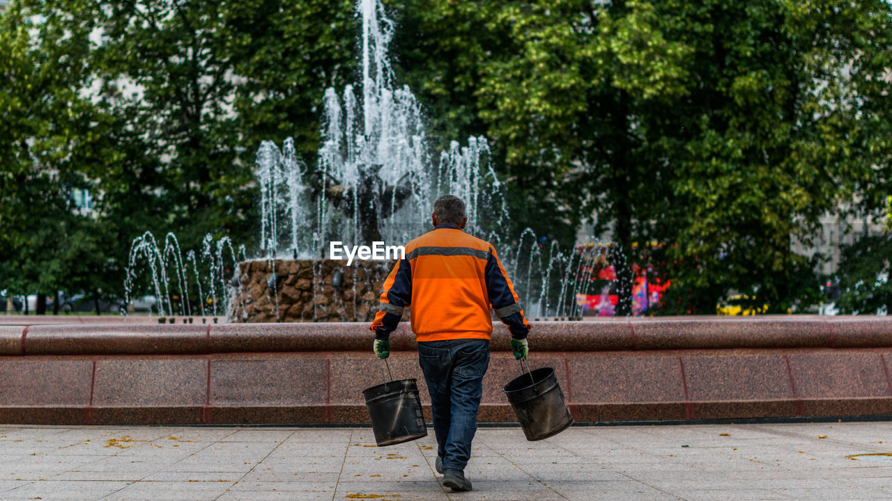 Rear view of male worker carrying buckets against fountain