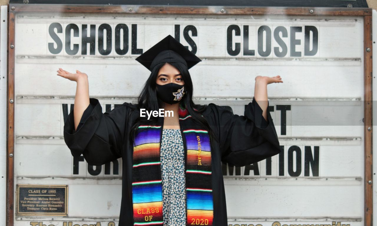 Portrait of young woman wearing mask and graduation gown standing against wall