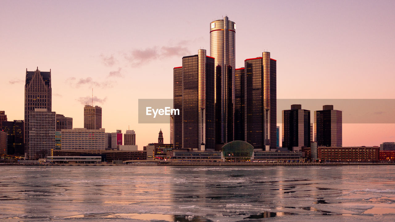 Modern buildings by frozen lake in city during sunset