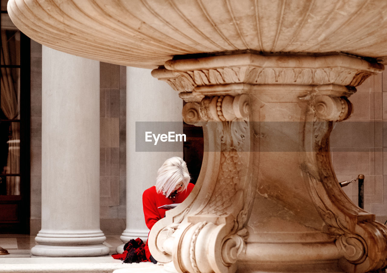 Lady in red reading below a large marble fountain.