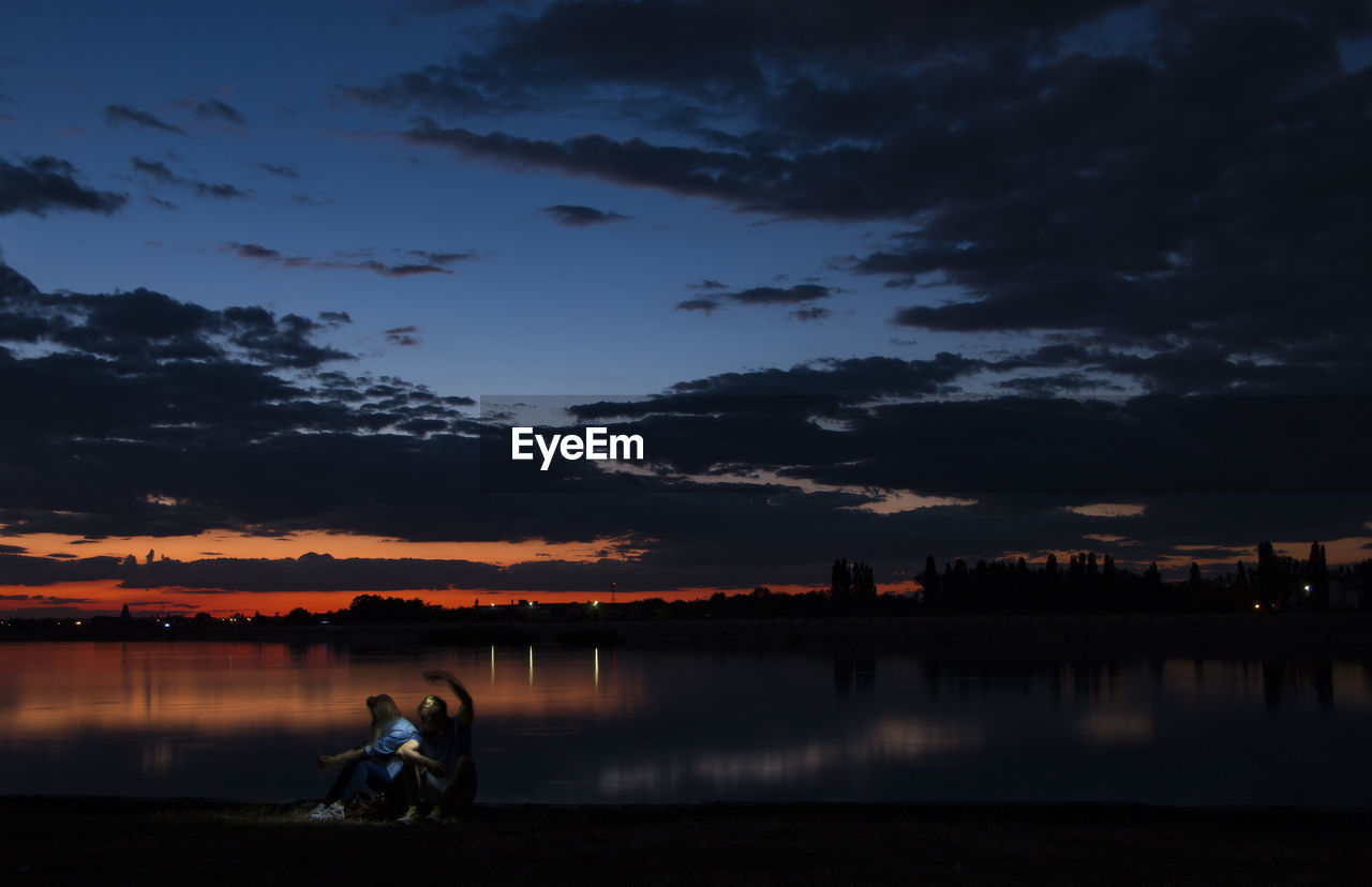 People sitting at lake against sky during sunset