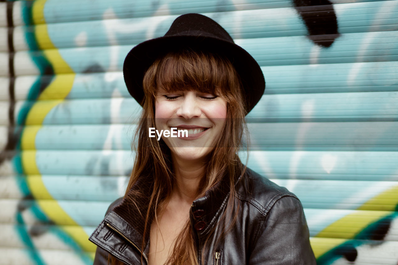 Smiling woman with eyes closed wearing hat against graffiti shutter in city