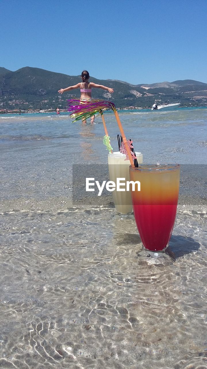 Drinking glasses on beach against girl standing in sea during sunny day