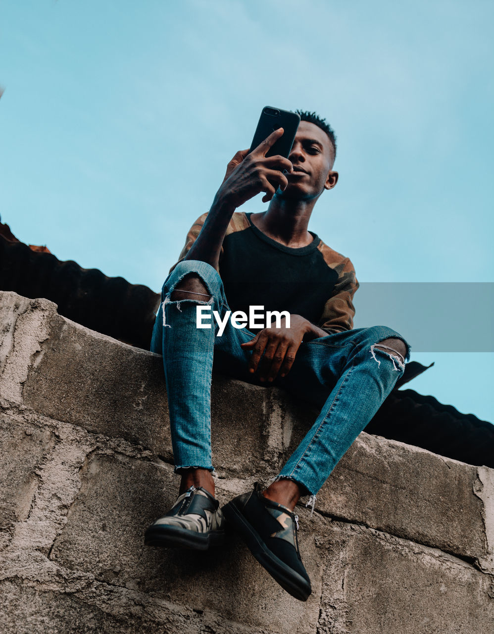 Full length of young man sitting on wall using a mobile phone against sky