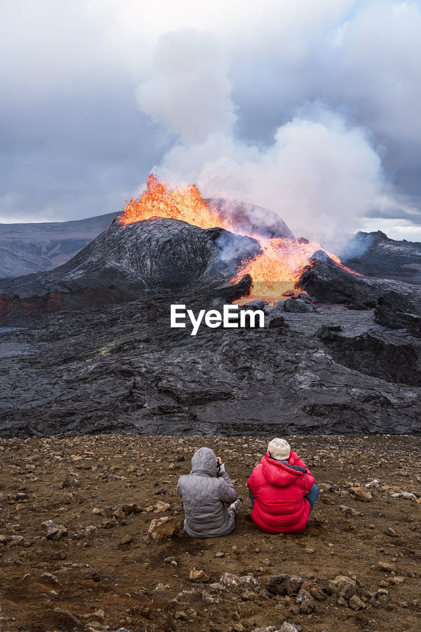 Back view of unrecognizable travelers admiring fagradalsfjall with fire and lava while taking photo and sitting on mount in iceland