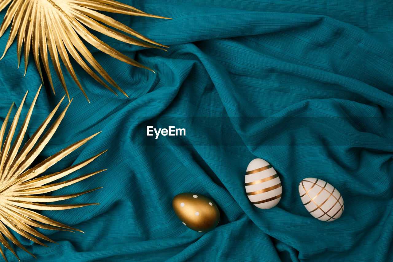 Minimal flat lay view of easter eggs and golden palm leaves against blue or cyan fabric background.