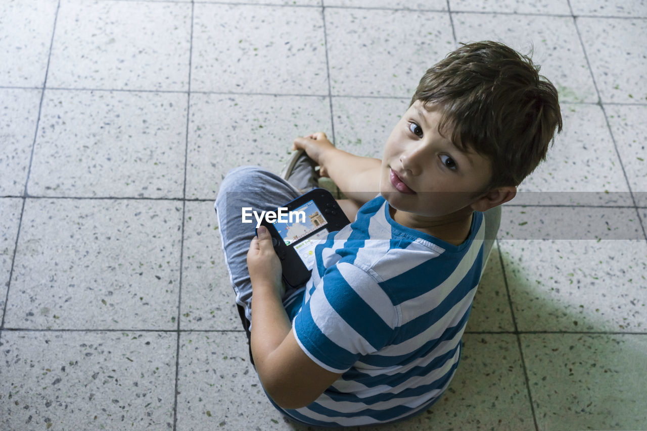 Portrait of cute boy sitting on tiled floor while holding video game at home