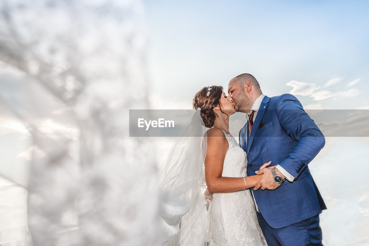 Low angle view of newlywed couple kissing against sky