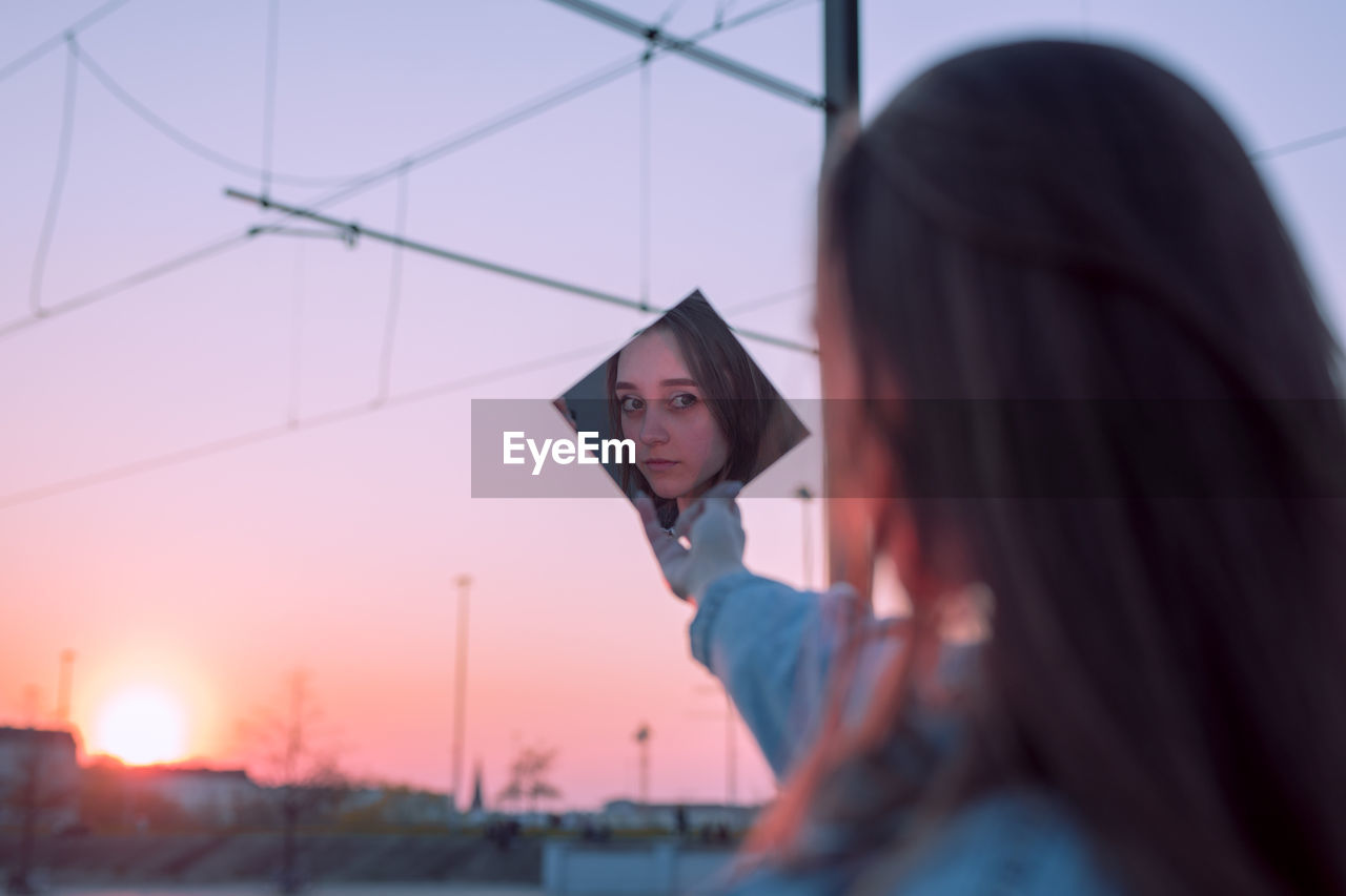 Woman holding mirror against sky during sunset