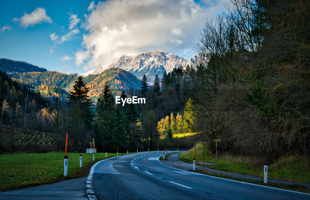 Empty road by trees and mountains against sky