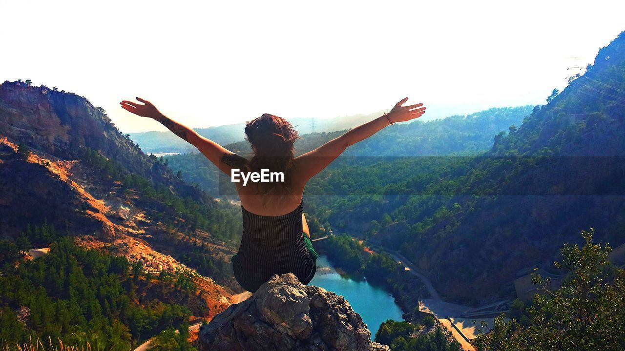 Rear view of woman with arms outstretched sitting on rock against mountain