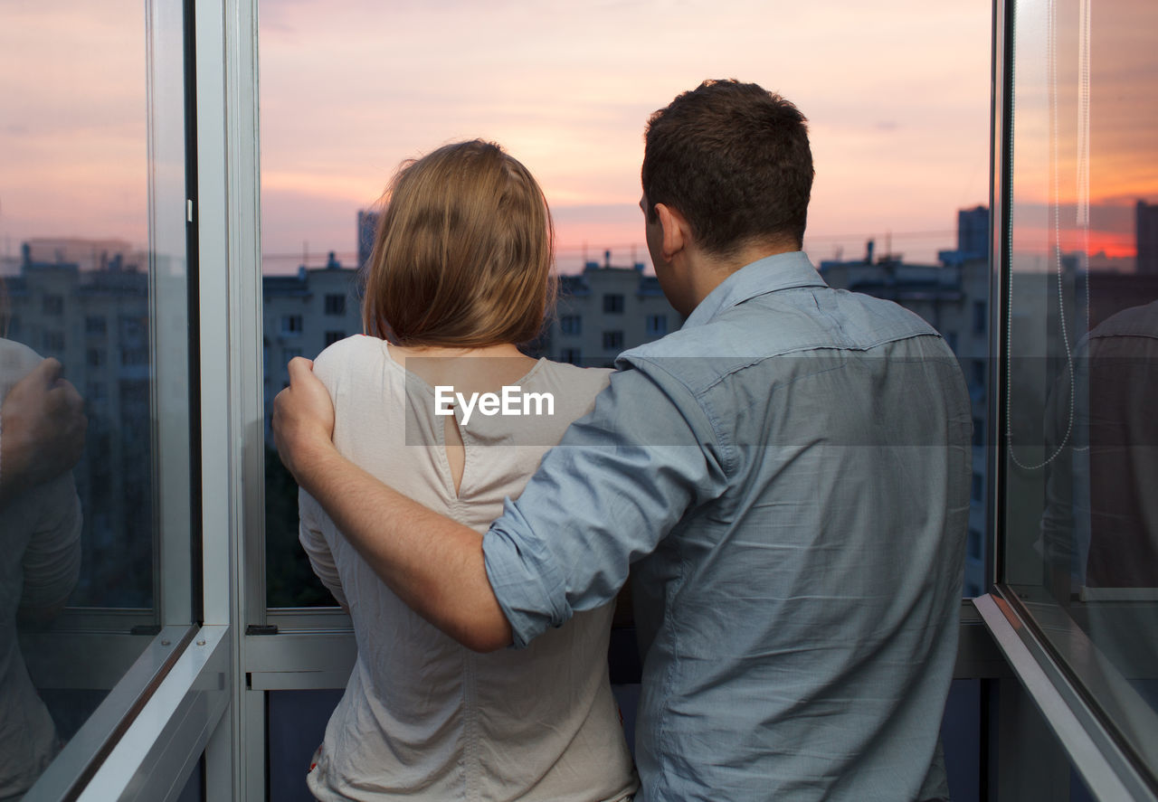 Rear view of man and woman looking through window during sunset