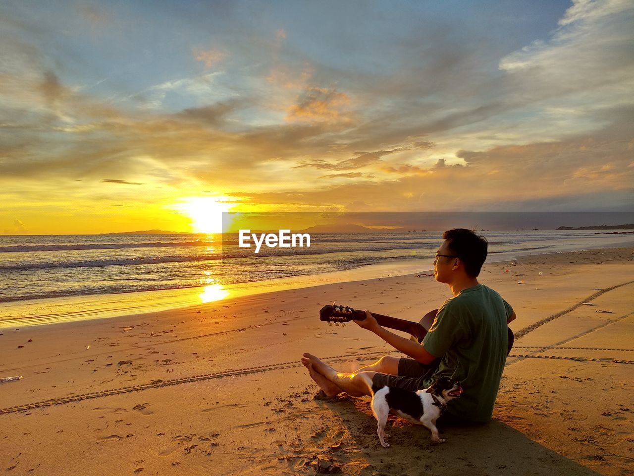 Full length of man playing guitar by dog at beach during sunset