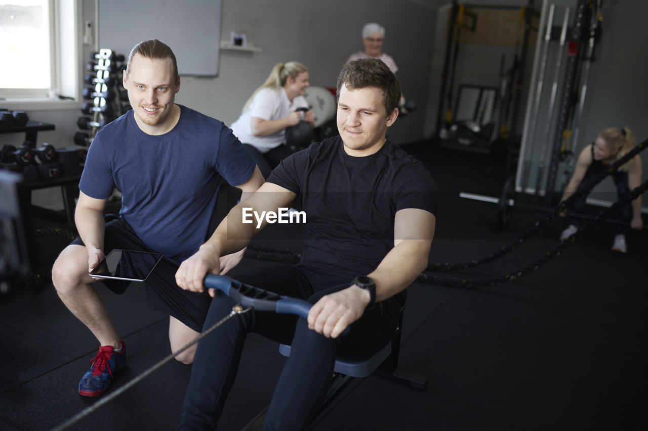 Smiling fitness instructor kneeling besides young man exercising on rowing machine at health club