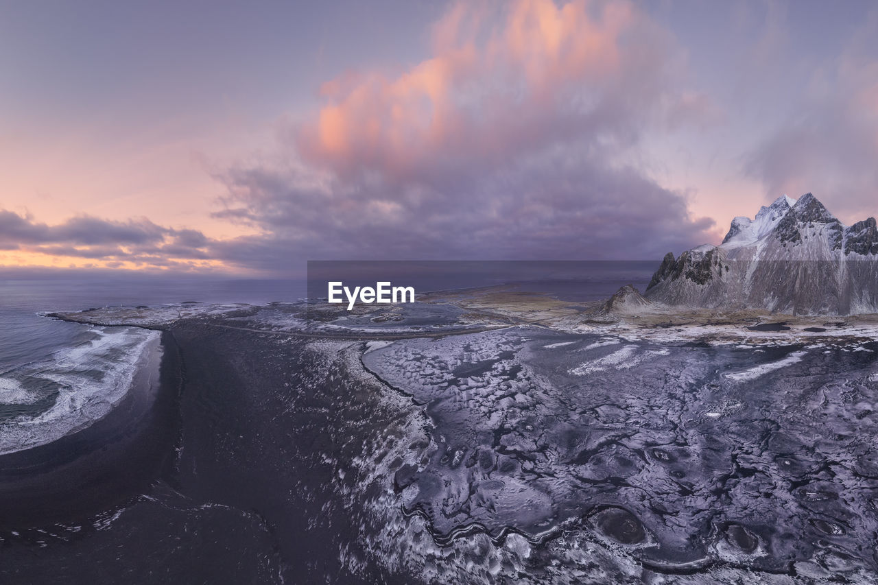 Drone view of calm frozen endless sea surrounding high rocky snowy vestrahorn mountain during picturesque sunset in stockness beach, iceland