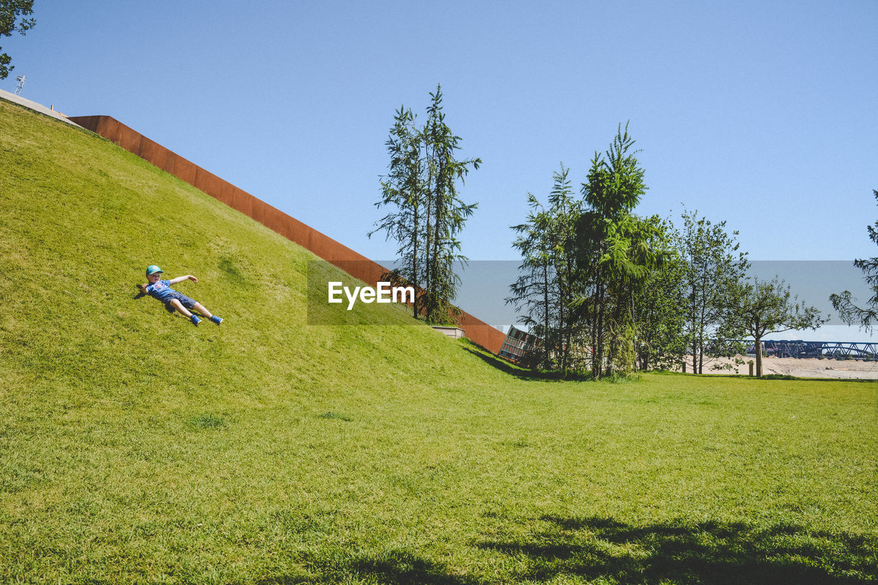Full length of boy lying on hill against clear sky during sunny day