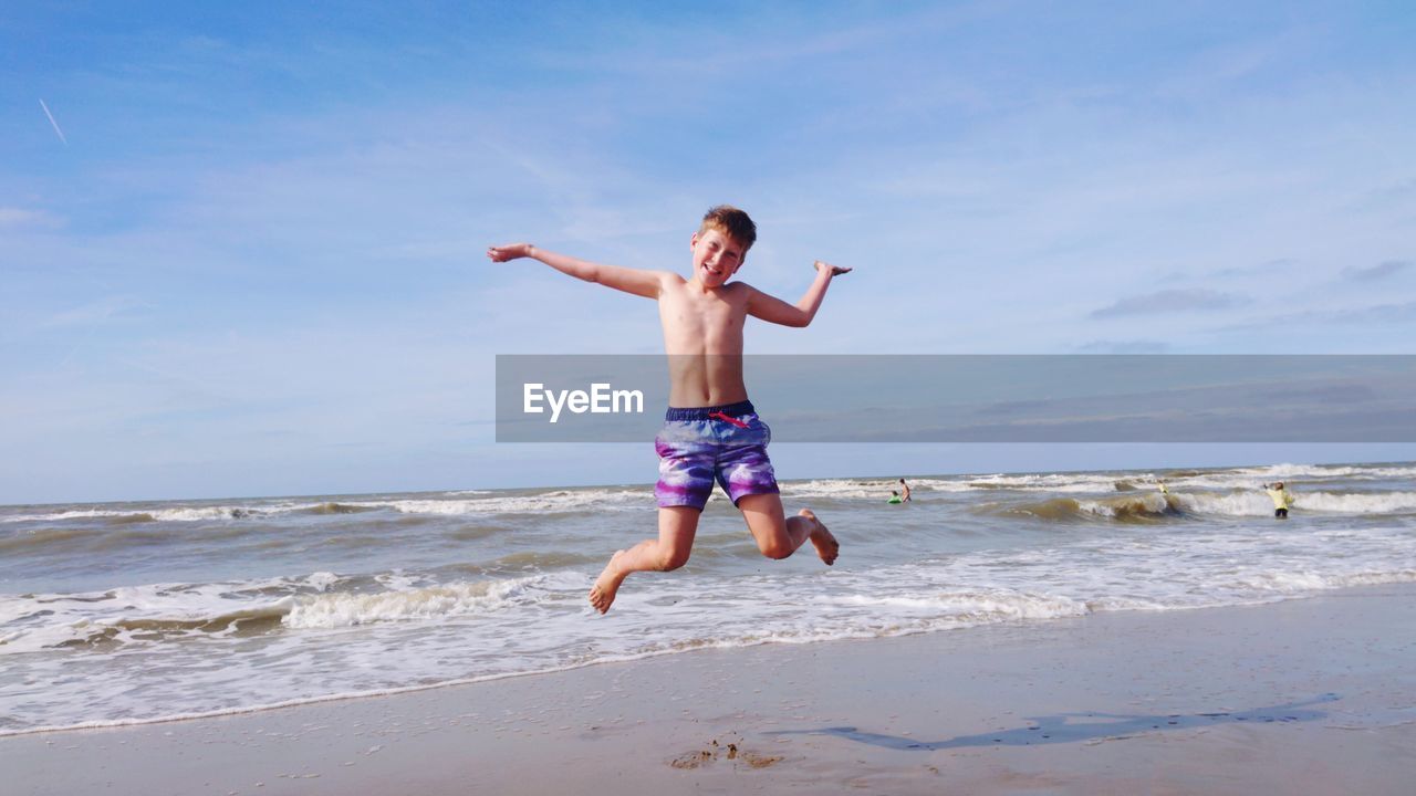 Full length portrait of shirtless boy with arms outstretched jumping at beach