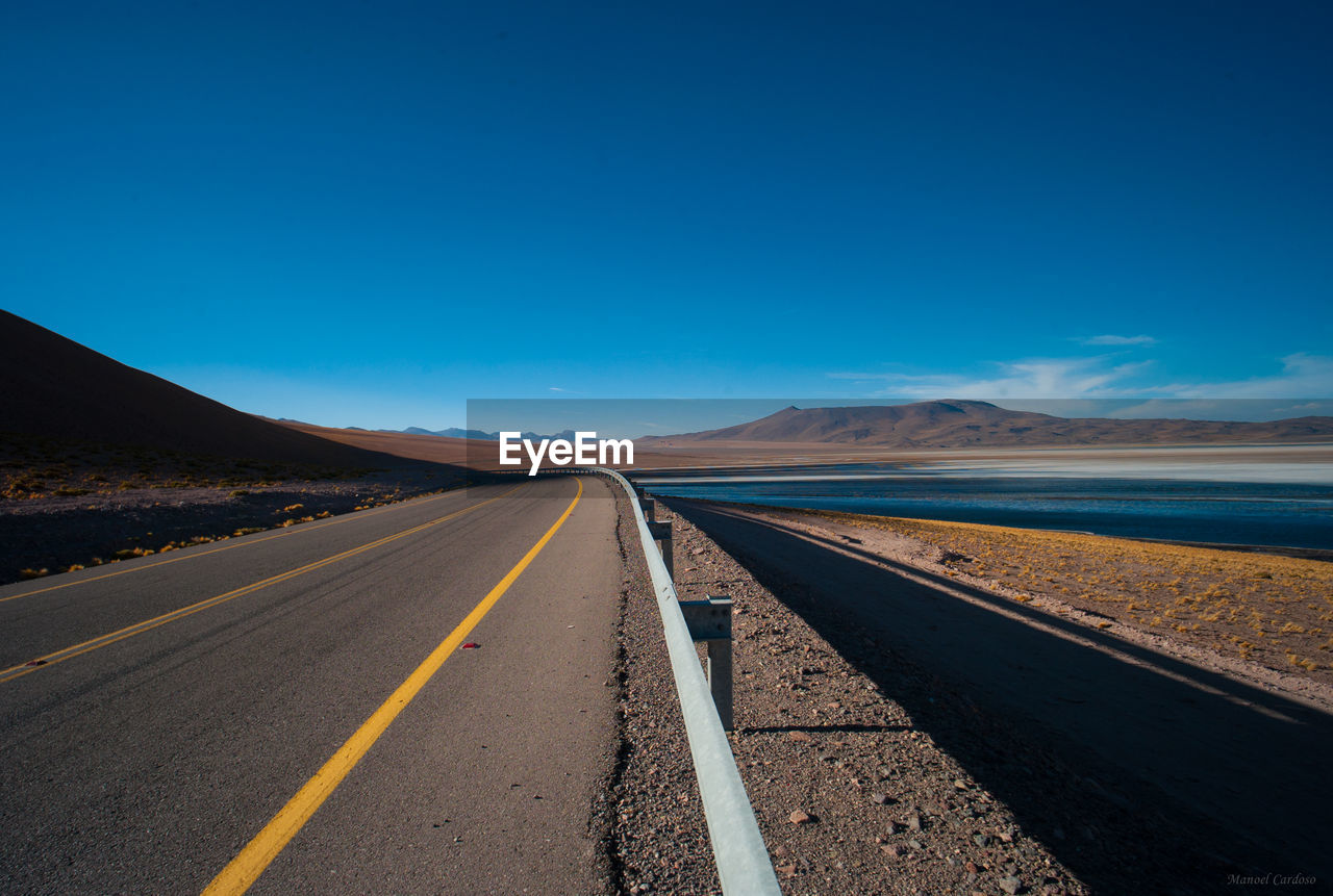 Empty road by lake against clear blue sky on sunny day