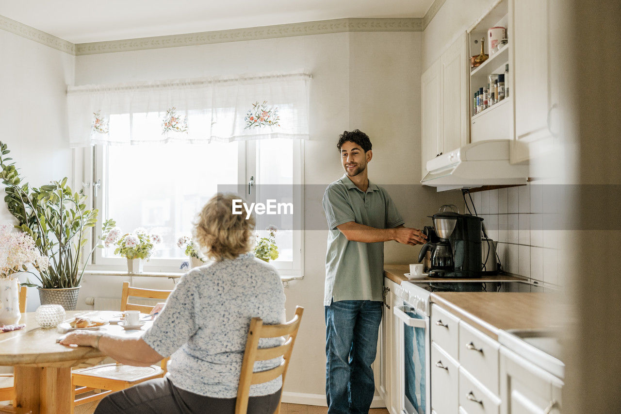 Male caregiver using coffee maker while talking with senior woman in kitchen at home