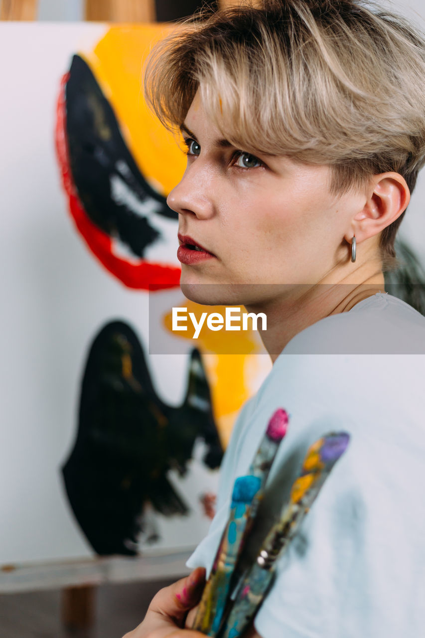 Creative portrait of beautiful young woman artist in studio with brushes.