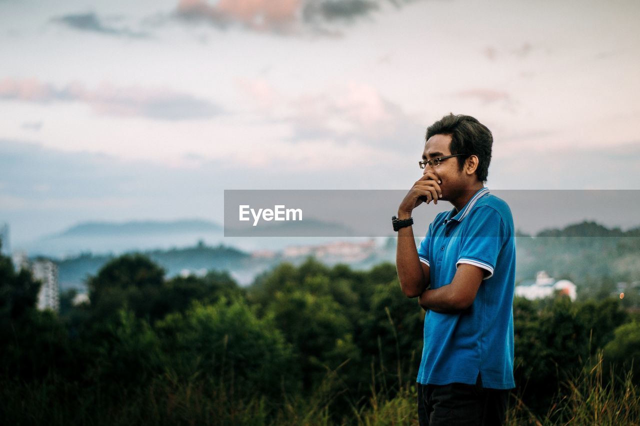 Young man looking away while standing in forest against sky