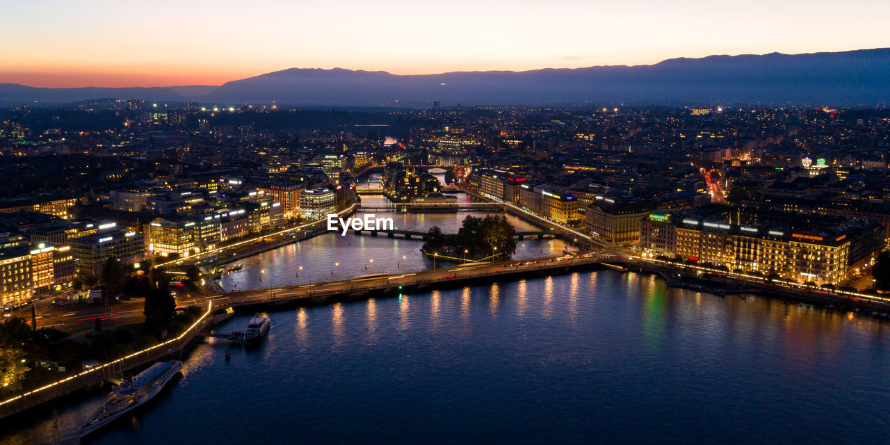 HIGH ANGLE VIEW OF ILLUMINATED CITY AGAINST SKY AT SUNSET