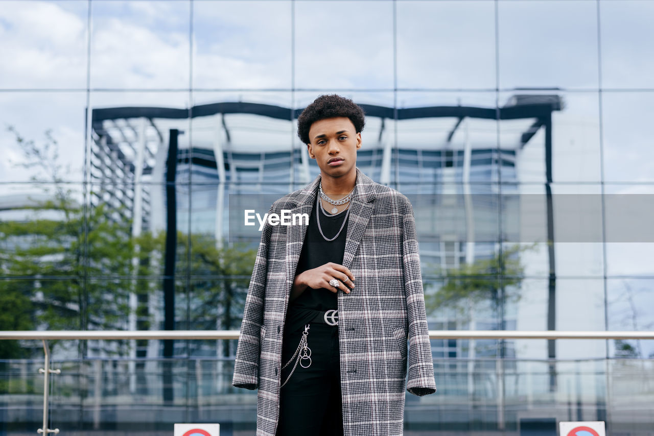 Confident young african american male teenager model in trendy tartan coat and stylish accessories looking at camera while standing against contemporary building with glass wall