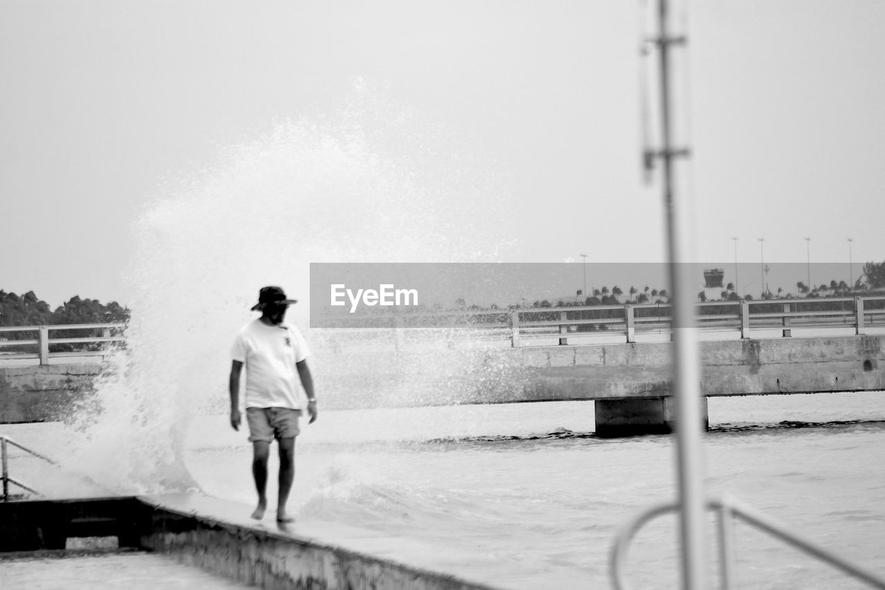 Man walking on retaining wall with wave splashing in sea