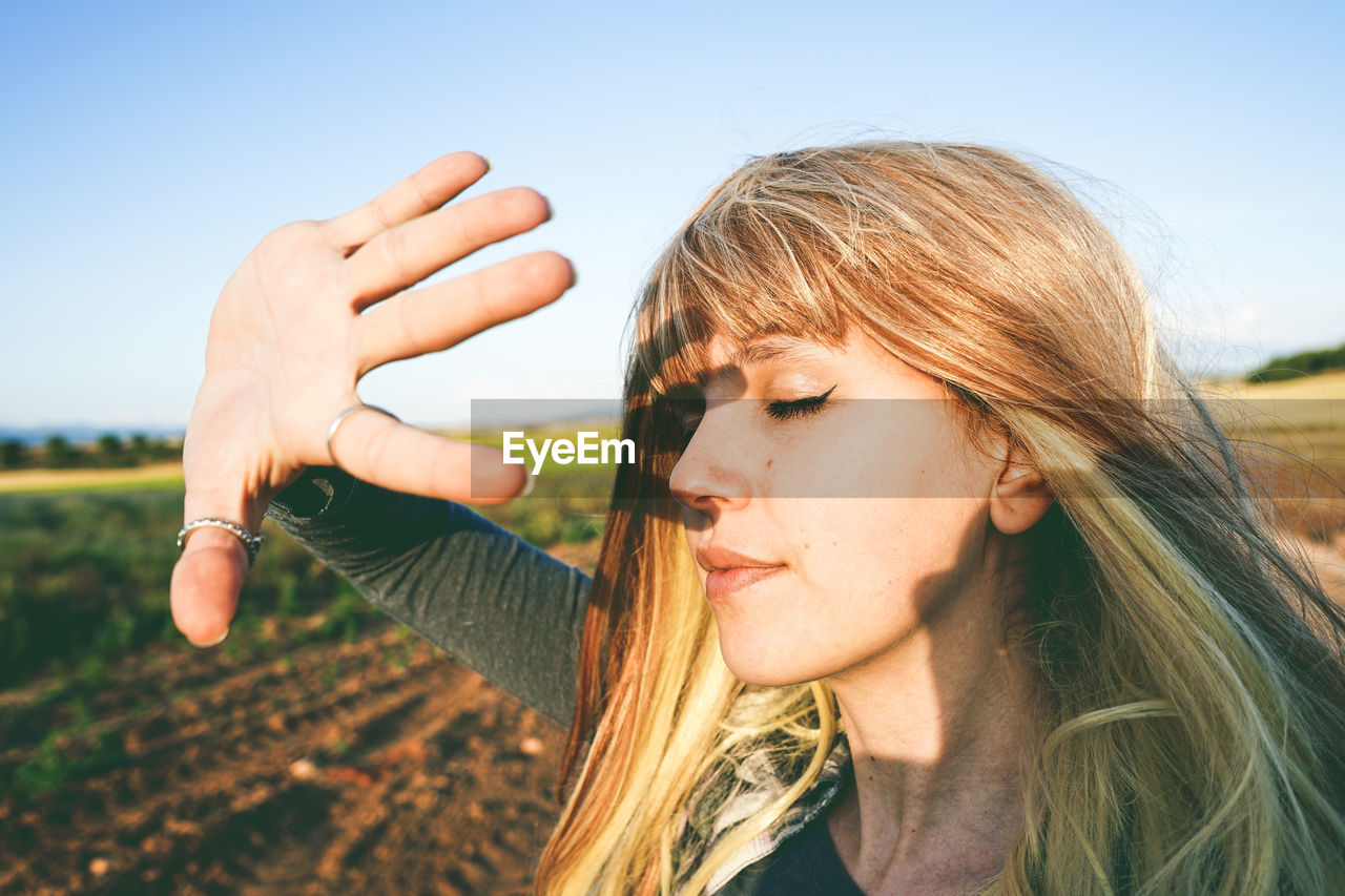 Young woman shielding eyes while standing at field against sky on sunny day