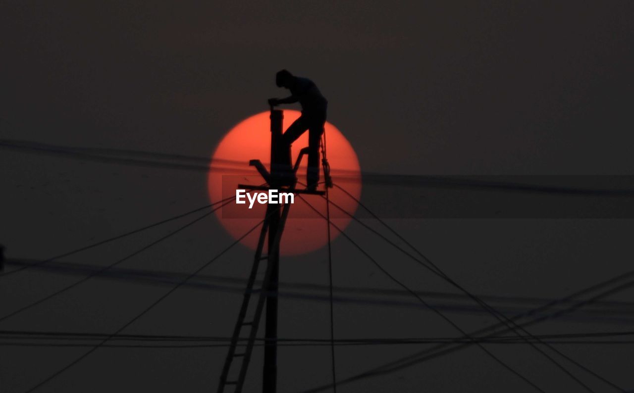 Low angle view of silhouette male electrician repairing power line against sun