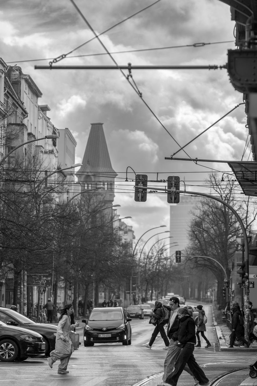 architecture, city, transportation, road, street, mode of transportation, black and white, car, built structure, building exterior, motor vehicle, urban area, monochrome, monochrome photography, city life, land vehicle, sky, infrastructure, city street, nature, cloud, cityscape, men, day, tree, vehicle, group of people, cable, adult, black, walking, sign, travel, building, outdoors, person, motion, crossing, downtown, traffic, wheel, electricity, full length, lifestyles, travel destinations, clothing