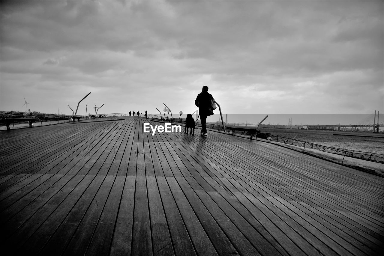 Full length of man with dog walking on boardwalk against sky